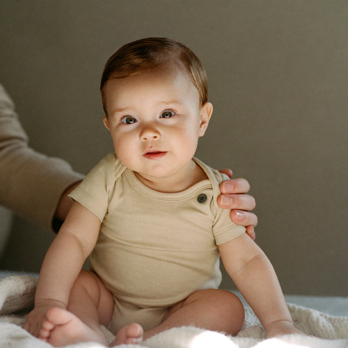 A baby sitting on a soft knit blanket with a parent's steadying hand